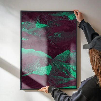 Person hanging a framed abstract mountain print with deep purple and vibrant green hues on a white wall.