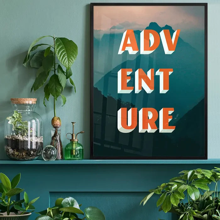 Bold "ADVENTURE" typography print with orange letters over teal mountain landscape, framed on a green shelf.