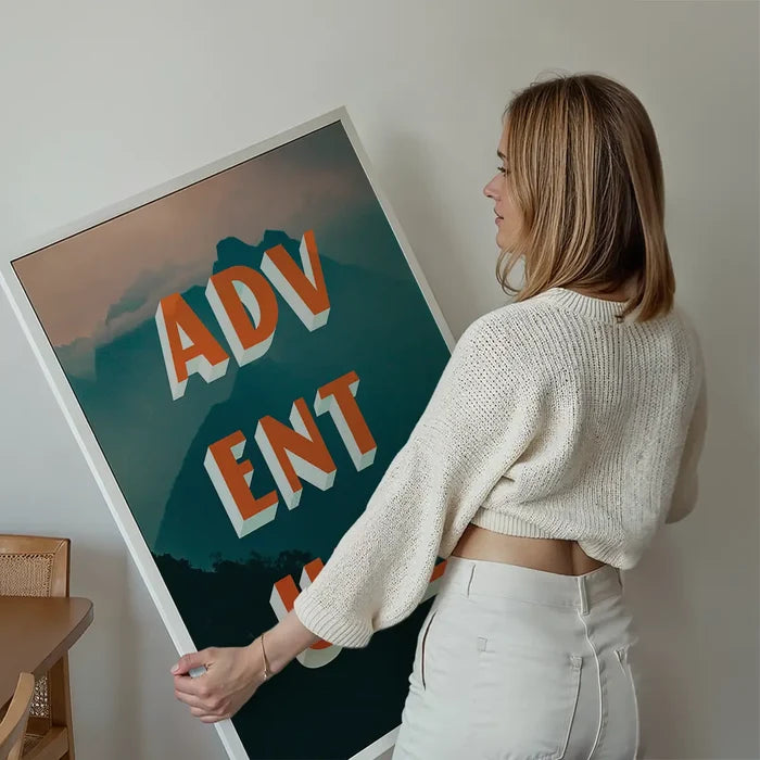Woman holding a framed "ADVENTURE" typography print with orange 3D letters over a teal mountain landscape.
