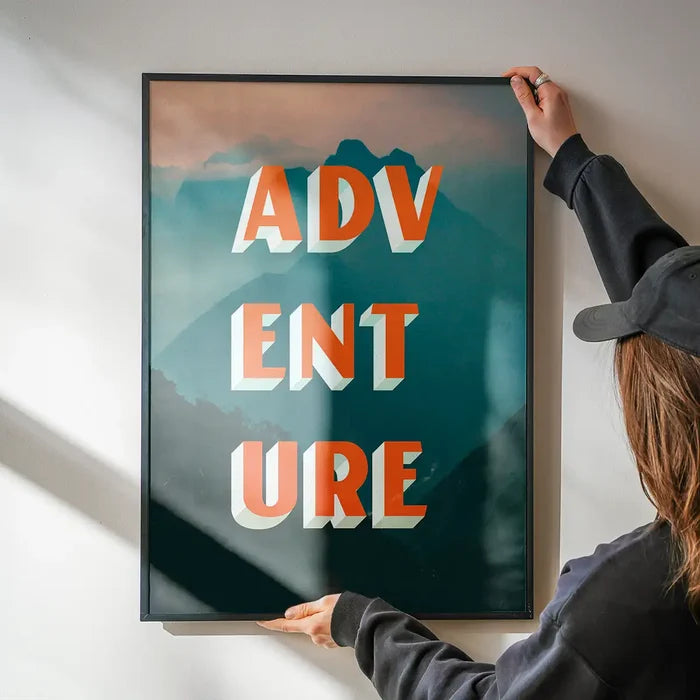 Framed "ADVENTURE" typography print with orange 3D letters over a teal and orange mountain landscape.