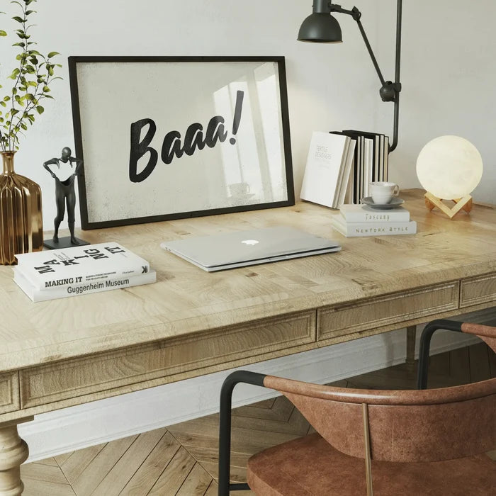 Minimalist "Baaa!" animal sound print in a black frame on a wooden desk with a laptop and books.