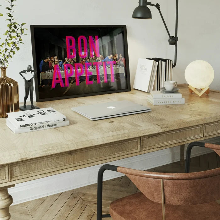 Black framed Bon Appetit typography print over a modern Last Supper painting on a wooden desk.