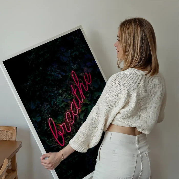 Woman holding a framed "breathe" neon sign print with pink script on a dark green leafy background.