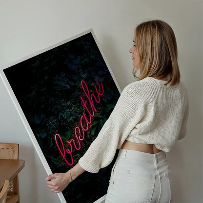 Woman holding a framed "breathe" neon sign print with pink script on a dark green leafy background.