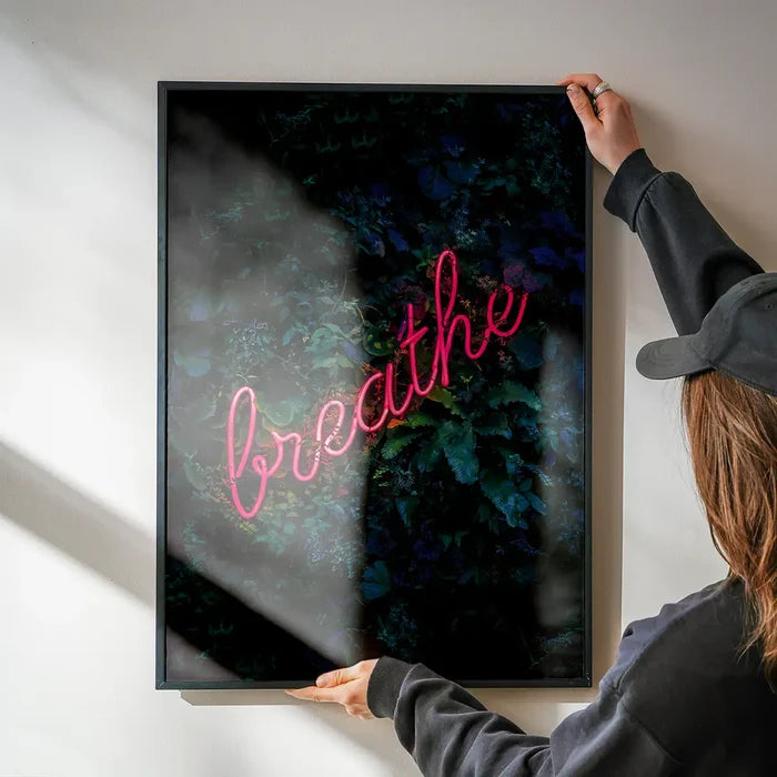 Framed "breathe" neon sign print on dark botanical background, held by hands against a white wall.