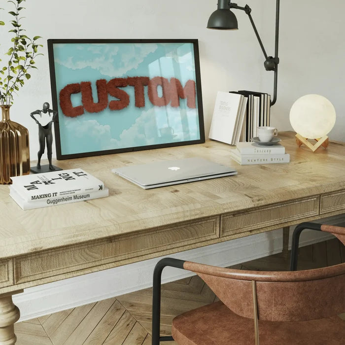 Brown fur-style "CUSTOM" text on a blue sky print in a black frame, on a wooden desk with a laptop.