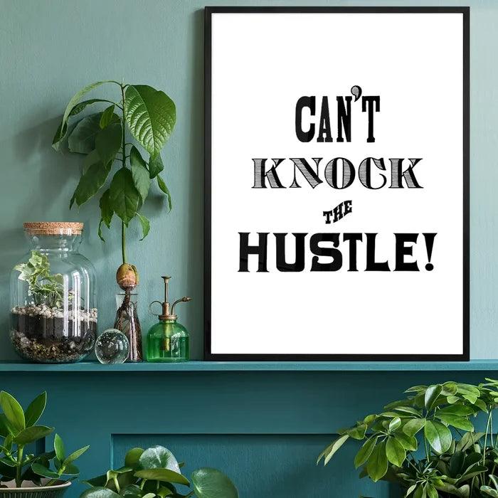 Black framed "Can't Knock The Hustle" typographic print on white paper, displayed on a teal shelf with plants.