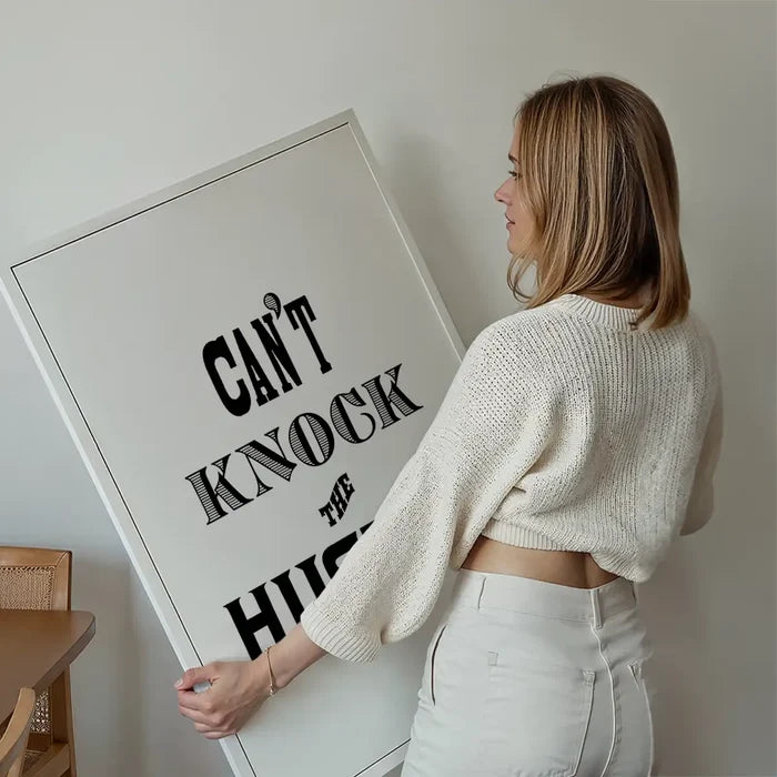 Woman holding a large white framed "Can't Knock The Hustle" typographic print with black text.