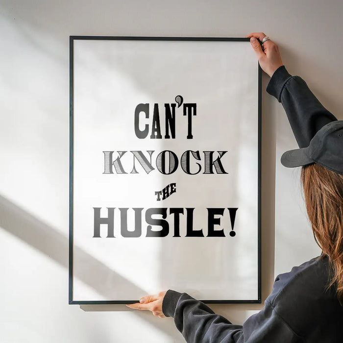 Black framed "Can't Knock The Hustle!" typographic print held by hands against a white wall.