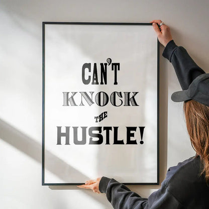 Black framed "Can't Knock The Hustle!" typographic print held by hands against a white wall.