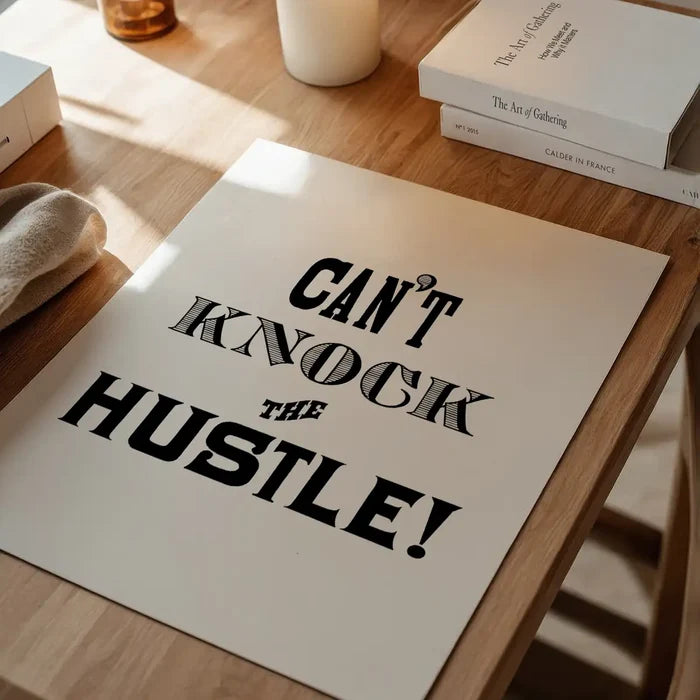 Can't Knock The Hustle" typographic print on white archival paper, black bold text, on a wooden table.