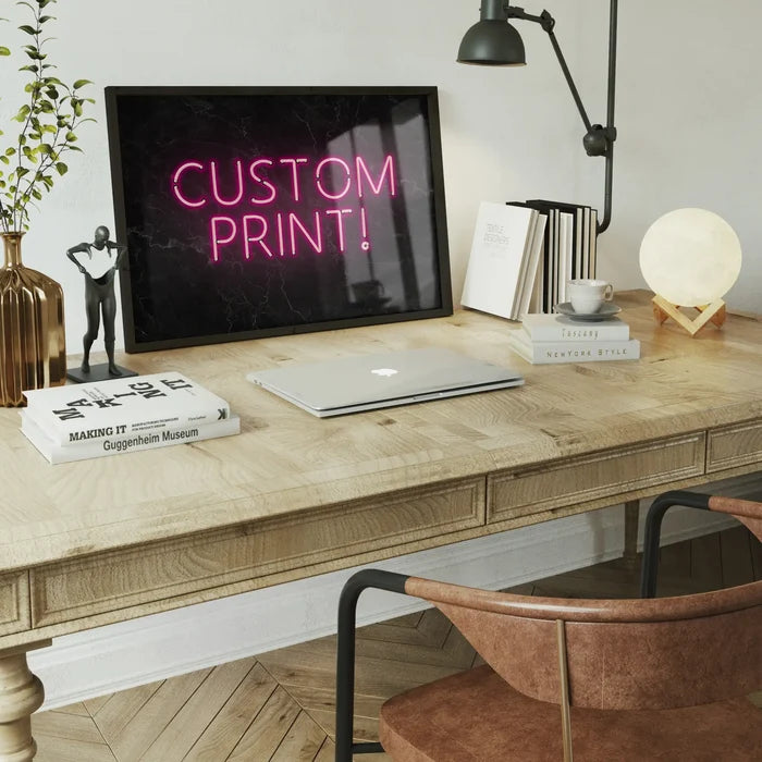 Pink "CUSTOM PRINT!" neon sign on black marble background, framed on a wooden desk with a laptop.
