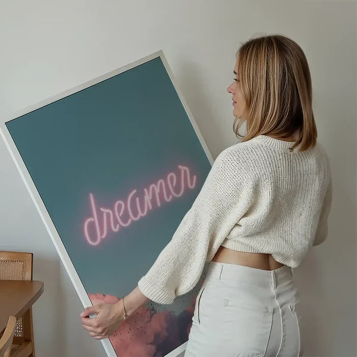 Woman holding a framed "dreamer" neon print with pink clouds on a blue sky background.