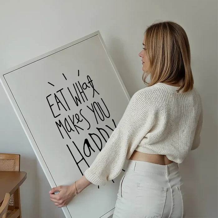 Woman holding a white framed typographic print with "Eat What Makes You Happy" in black script.