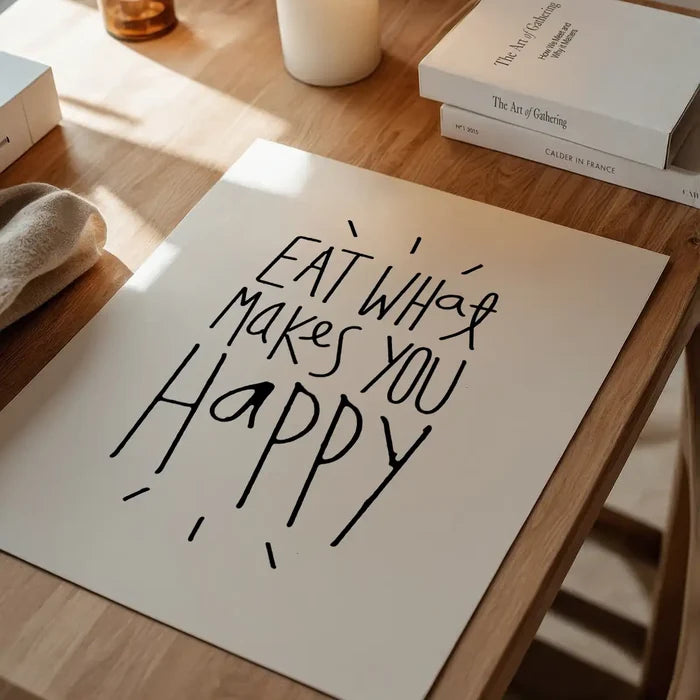 White typographic print with "Eat What Makes You Happy" in black, on a wooden table with books and candles.