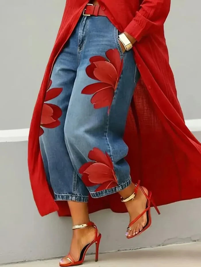 A person wearing blue cropped jeans with large red floral applique patches, a flowing red cardigan, and red high-heeled sandals, standing against a light gray wall.