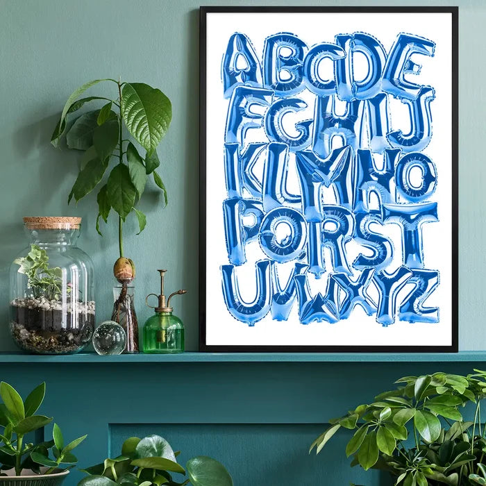 Blue foil balloon alphabet A-Z educational print in a black frame, displayed on a teal shelf with plants.
