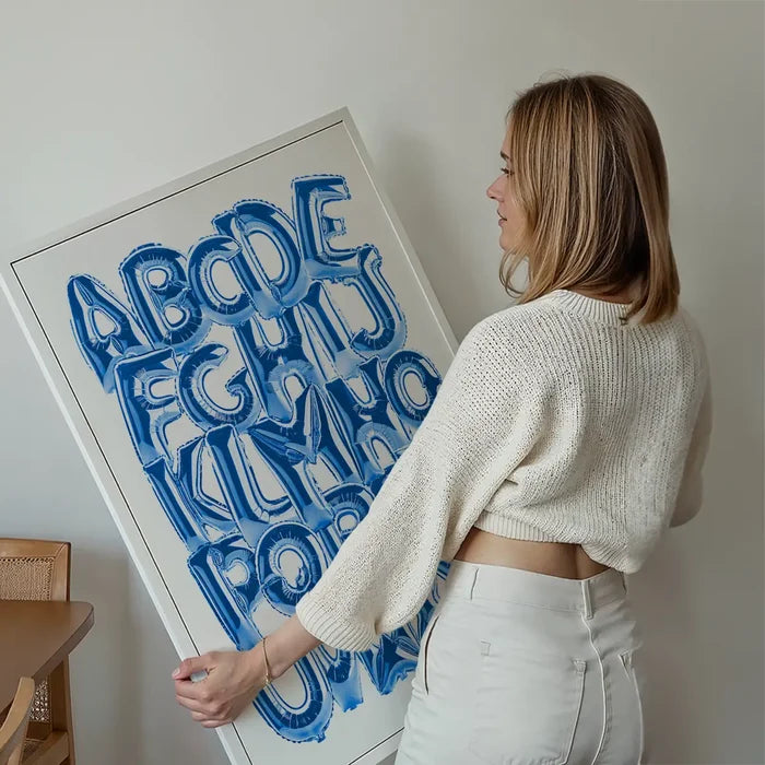 Woman holding a large white framed poster with blue foil balloon-style alphabet letters A-Z.