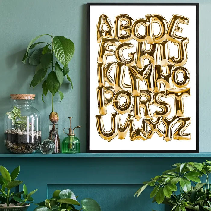Gold foil balloon alphabet A-Z educational print in a black frame, on a teal shelf with plants.
