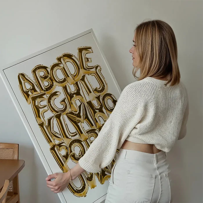 Woman holding a framed educational poster with gold foil balloon alphabet letters A-Z on a white background.