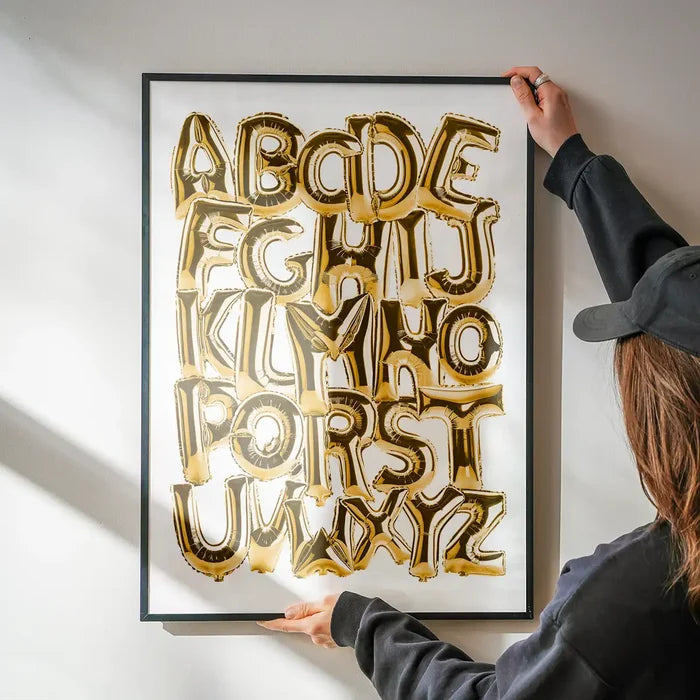 Gold foil balloon alphabet A-Z educational print in a black frame, held by hands on a white wall.