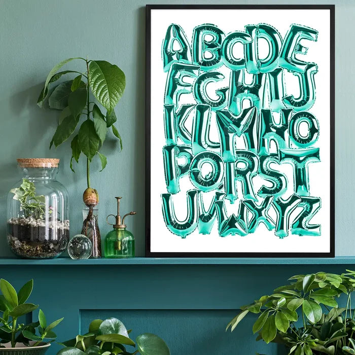 Teal foil balloon alphabet A-Z educational print in a black frame, styled on a green shelf.