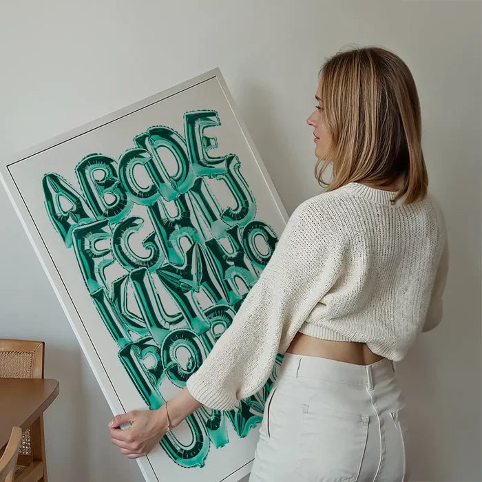 Woman holding a framed educational poster with teal foil balloon alphabet letters A-Z on a white background.