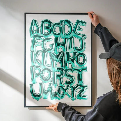 Framed teal foil balloon alphabet print (A-Z) being hung on a white wall.