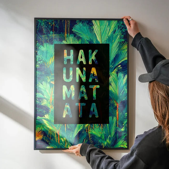 Hakuna Matata tropical print with green palm leaves and black frame, held by hands on a white wall.