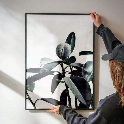 Person hanging a framed "Leaves Print" art piece with subtle green natural colors on a white wall.