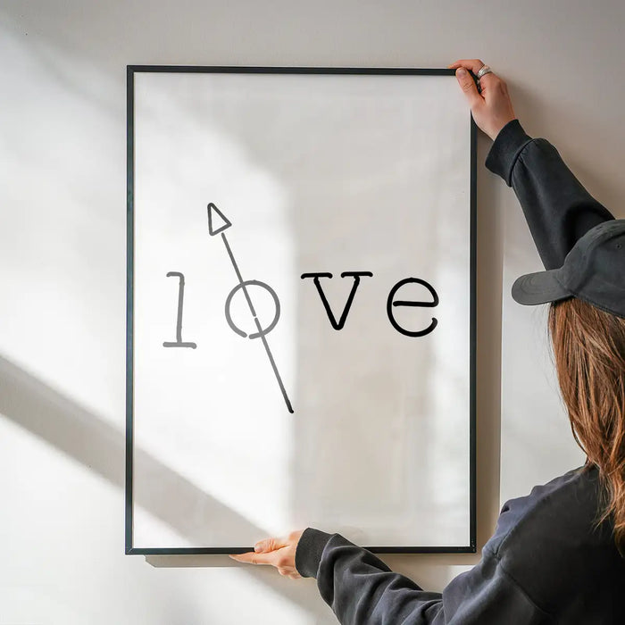Black framed "love" typography print with a crossed-out "o" and arrow, held against a white wall.