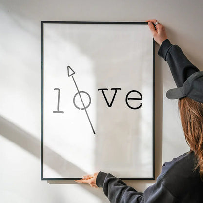 Black framed "love" typography print with a crossed-out "o" and arrow, held against a white wall.