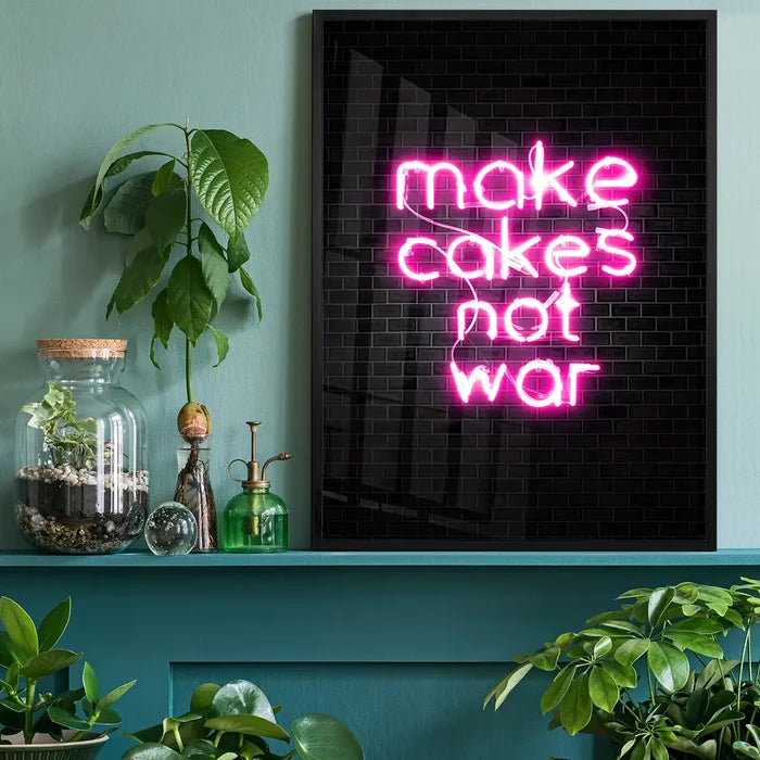 Pink neon "make cakes not war" print on black metro tiling, framed and displayed on a green shelf with plants.