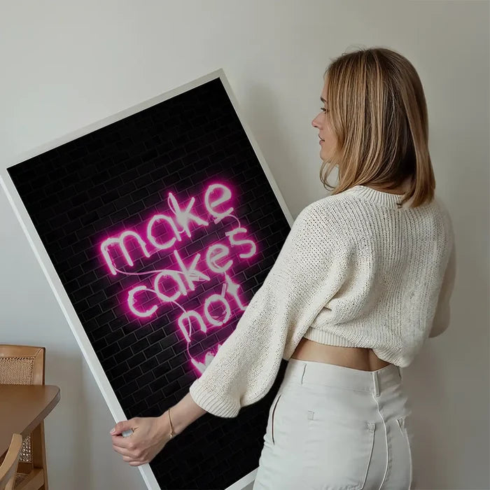 Woman holding a framed "make cakes not war" pink neon print on black metro tile background.