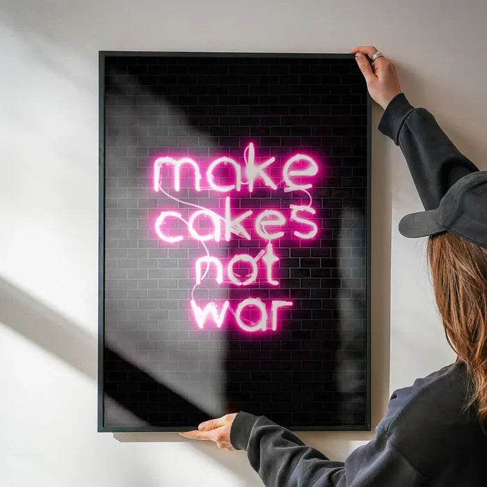 Framed "make cakes not war" neon pink print on black metro tile background, held by hands.