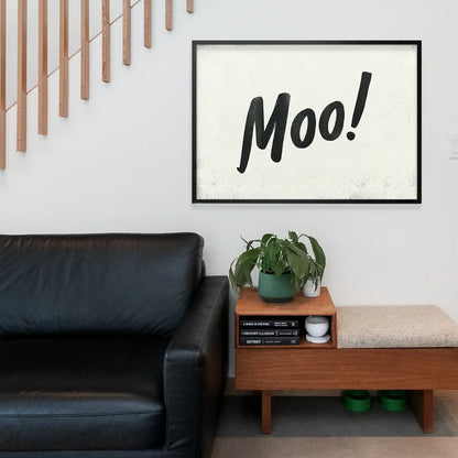 Black framed "Moo!" brush script print on textured off-white paper, above a black leather sofa.