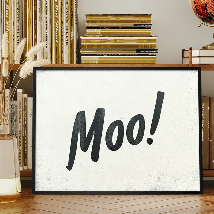 Black-framed "Moo!" print with hand-painted brush script typography on a textured white background, displayed on a wooden shelf.