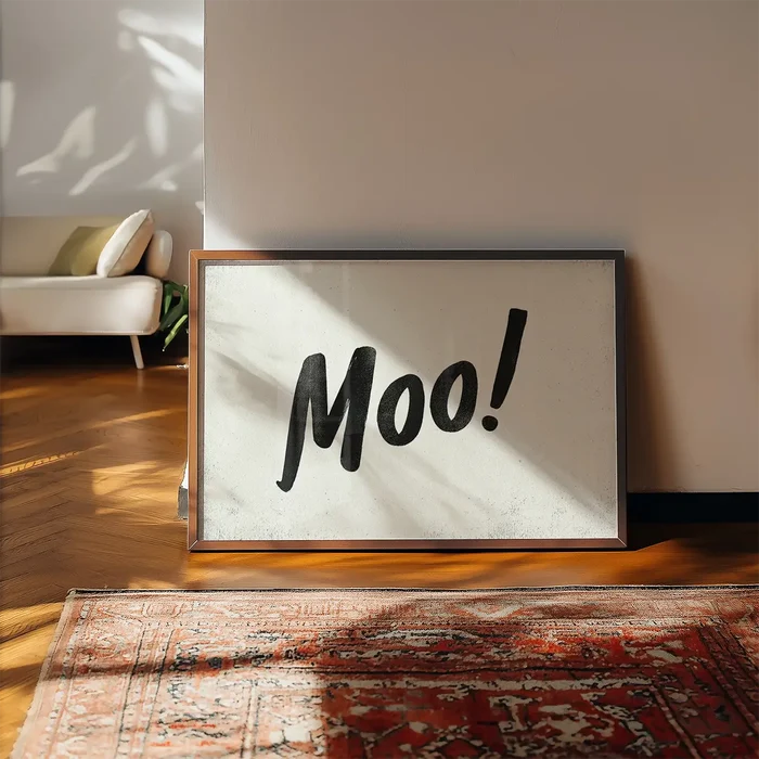 Framed "Moo!" print with black brush script on a light background, leaning against a wall above a patterned rug.