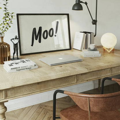 Framed "Moo!" brush script print on a wooden desk with a laptop, books, and decorative items.