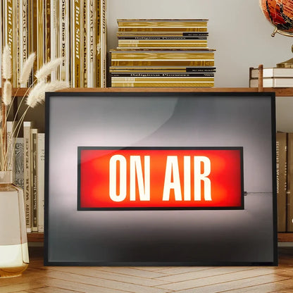 Framed retro "ON AIR" lightbox print with a glowing red sign, on a wooden shelf with books.