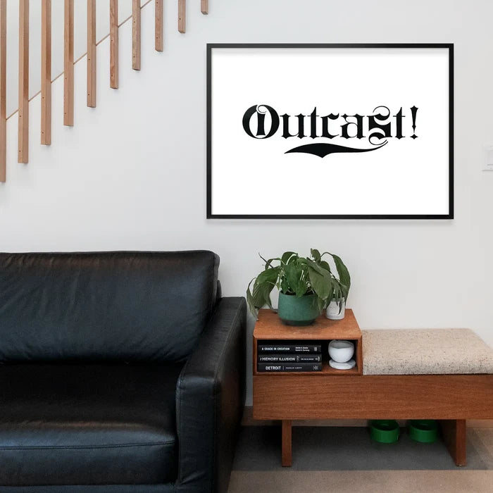 Black framed "Outcast!" typography print in a modern living room above a black leather sofa and wooden bench.