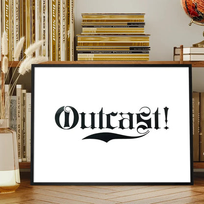 Black framed "Outcast!" typography print with ornate black lettering on white paper, displayed on a wooden shelf.