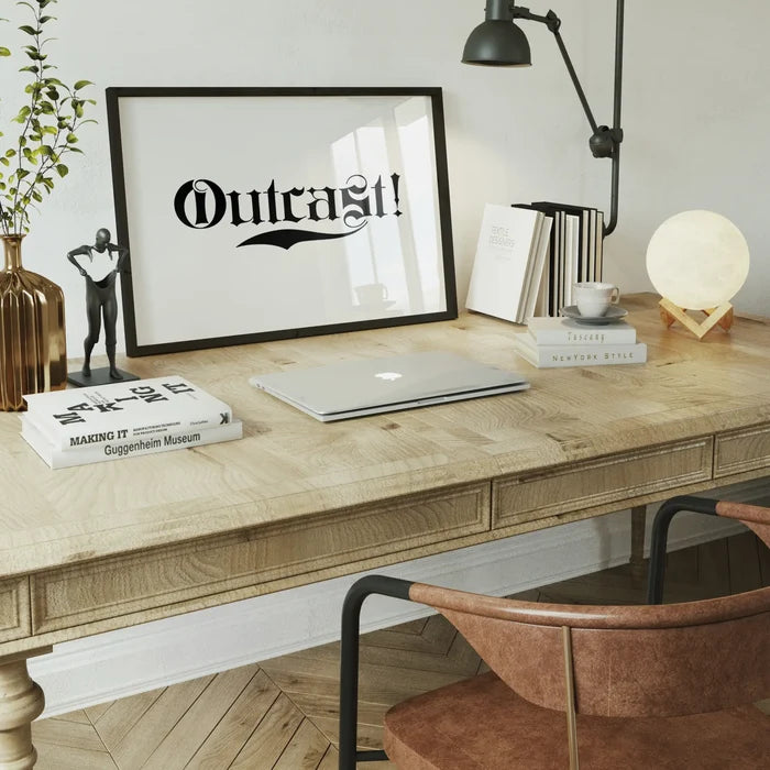Black framed "Outcast!" typography print on a light wood desk with a laptop, books, and a moon lamp.