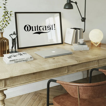 Black framed "Outcast!" typography print on a light wood desk with a laptop, books, and a moon lamp.