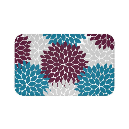 Rectangular mat rug with rounded corners, featuring a bold, graphic floral pattern. The pattern consists of large, stylized flowers reminiscent of dahlias or chrysanthemums. These flowers are made up of numerous pointed, overlapping petal shapes. The overall look is modern and graphic, with the contrasting jewel tones (maroon, gray, and teal flower petals) set against a white background.