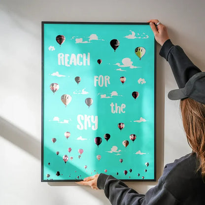 Teal "Reach For The Sky" print with colorful hot air balloons in a black frame, held by hands.