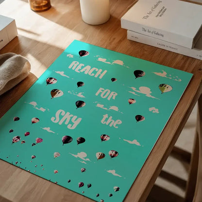 Teal print with "Reach For The Sky" text, hot air balloons, and clouds on a wooden table.