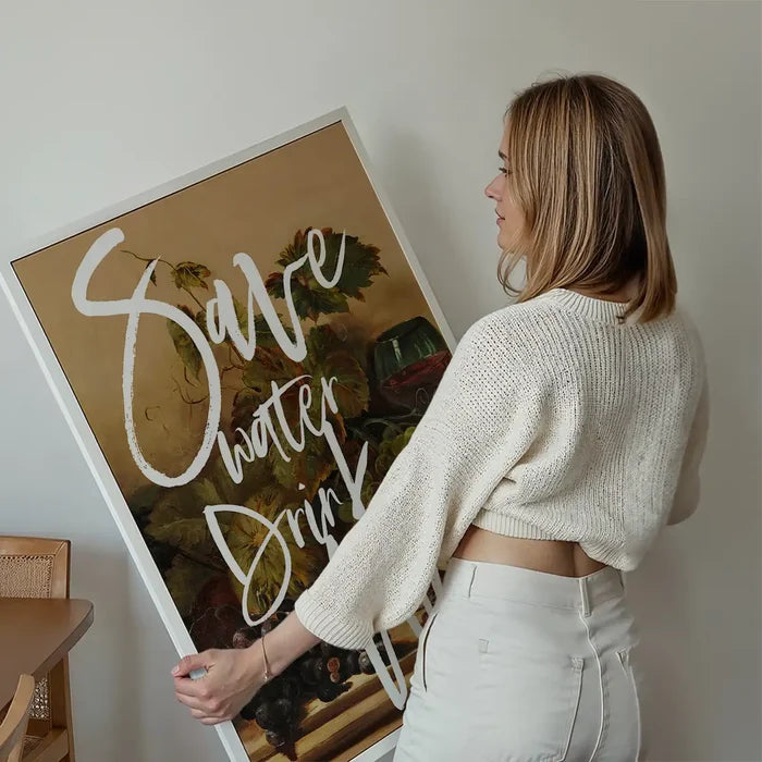 Woman holding a framed "Save Water Drink Wine" typography print with a vintage still life background.