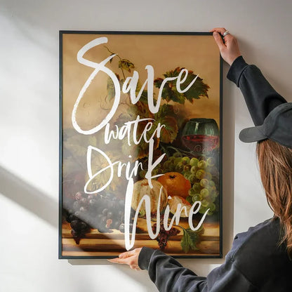 Framed "Save Water Drink Wine" typography print on a still life background, held by hands.