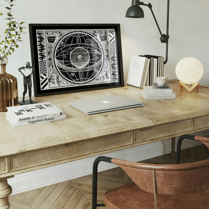 Black framed Sceno Systematis Astrology print on a light wood desk with a laptop and books.